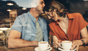 Two adults in a coffee shop. The man is kissing the lady's head